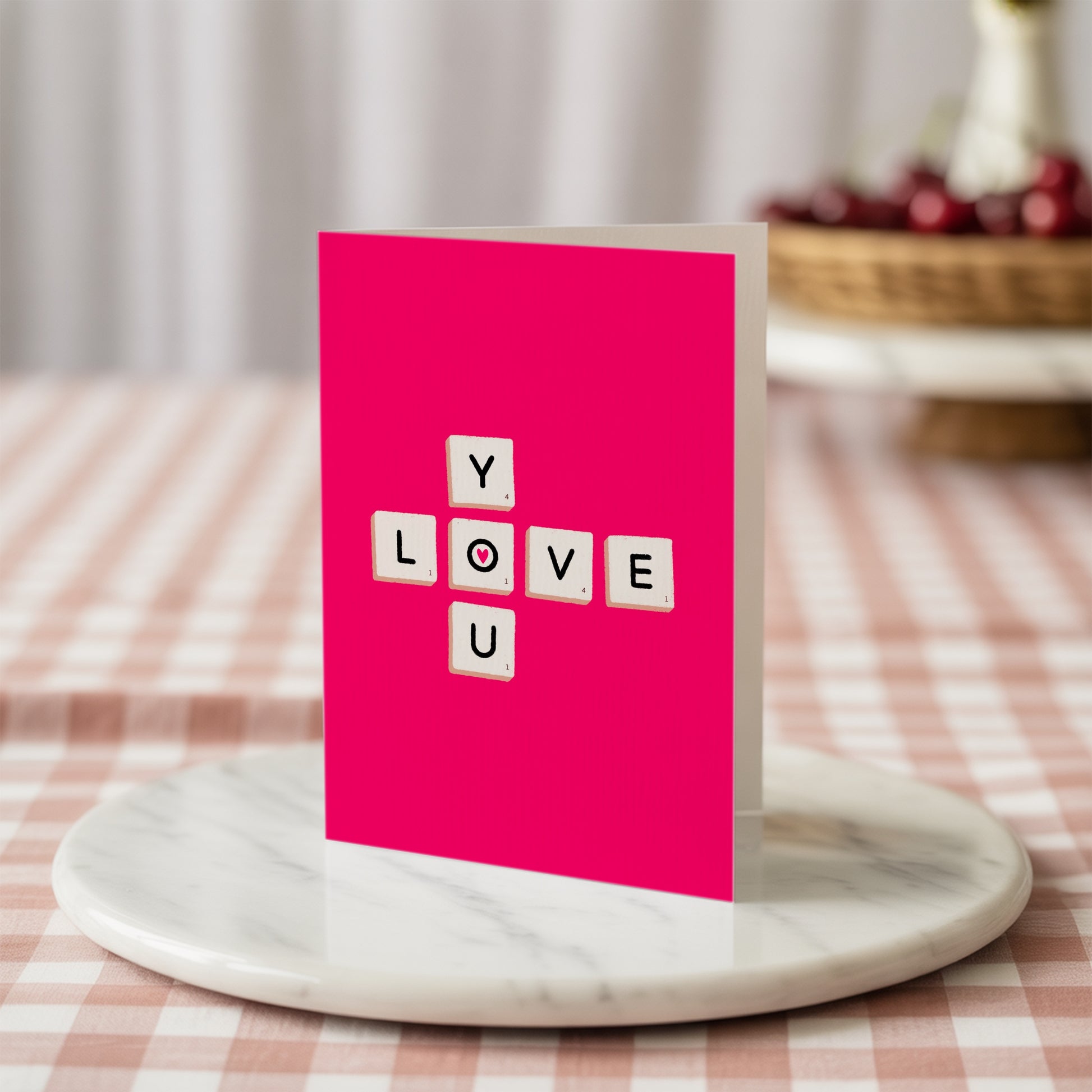 Pink card with 'LOVE YOU' in scrabble tiles on a marble stand with a checkered tablecloth background.
