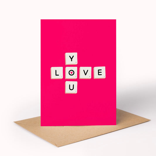 A bright pink greeting card standing upright on a brown kraft envelope against a clean white background. The card features white letter tiles spelling LOVE, with the letter O styled like a red heart.