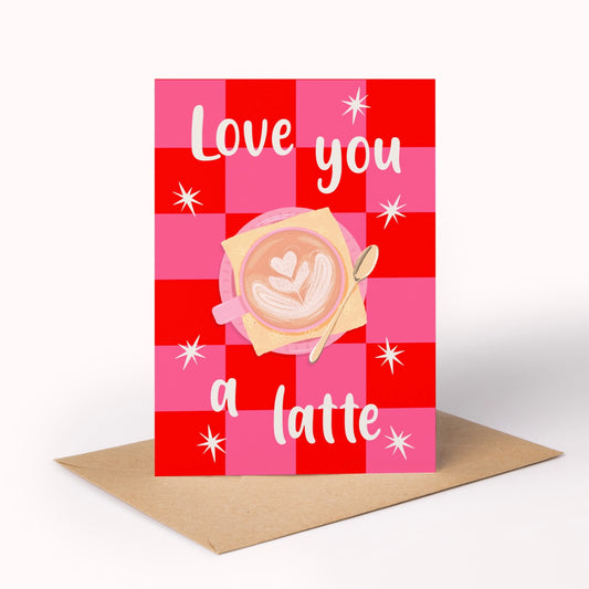 A greeting card with a red and pink checkered pattern, featuring the text 'Love you a latte' in white script around an illustration of a cup of latte art, displayed on a brown envelope against a white background.
