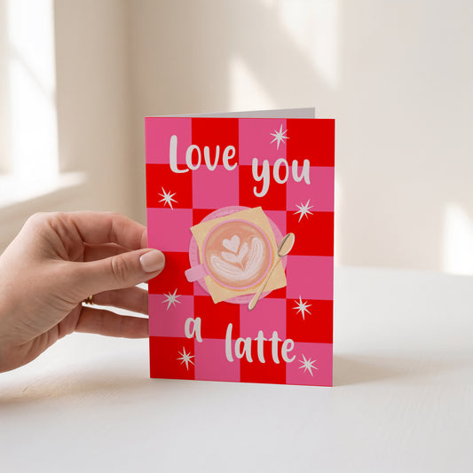 A hand holds a greeting card with a pink and red checkered pattern, featuring the text 'Love you a latte' and an illustration of a latte art coffee cup.