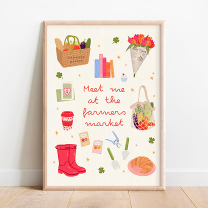 A framed art print featuring a whimsical 'Meet me at the farmers market' illustration, leaning against a white wall on a light wood floor.