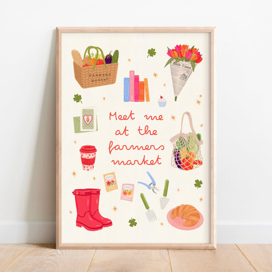 A framed art print featuring a whimsical 'Meet me at the farmers market' illustration, leaning against a white wall on a light wood floor.