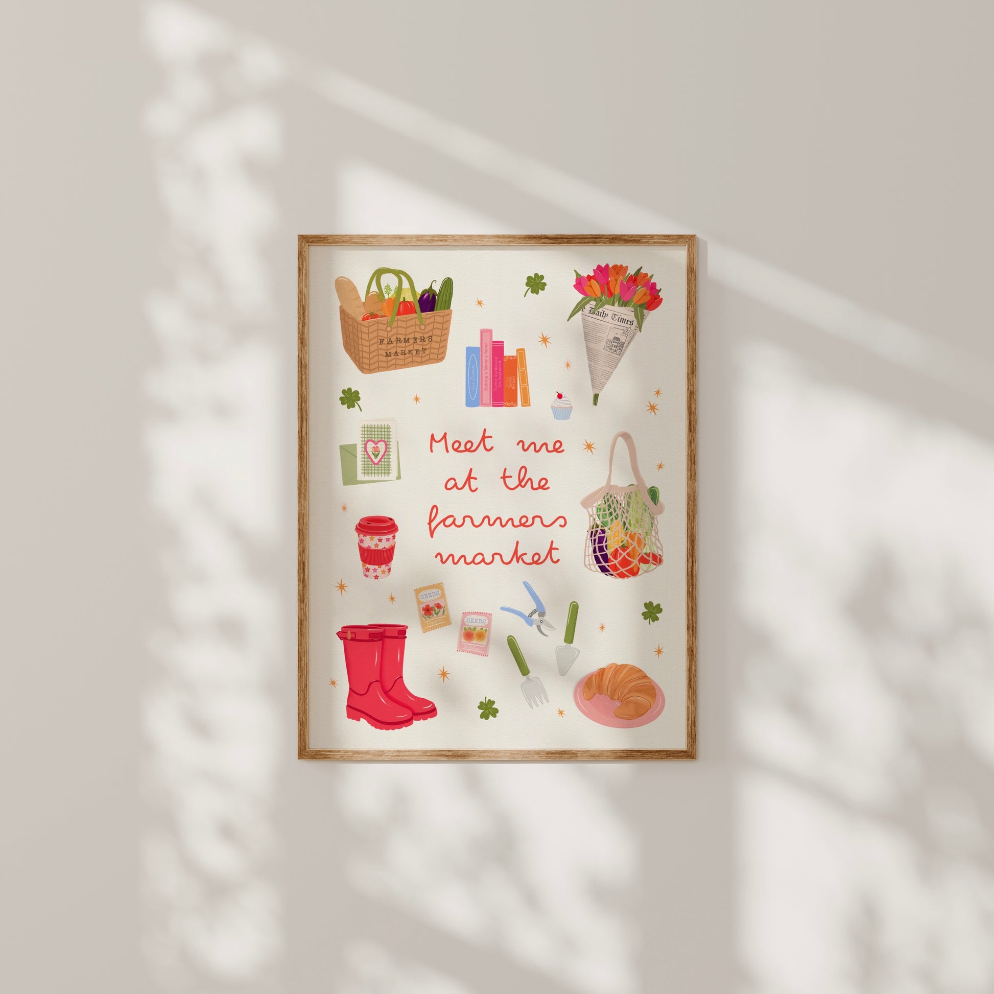 A framed art print featuring a whimsical autumn farmer's market illustration with the text 'Meet me at the farmers market' in red script. The print includes colorful drawings of a basket of vegetables, books, flowers, a tote bag, a red rain boot, gardening tools, and a plate of food, all set against a cream background.