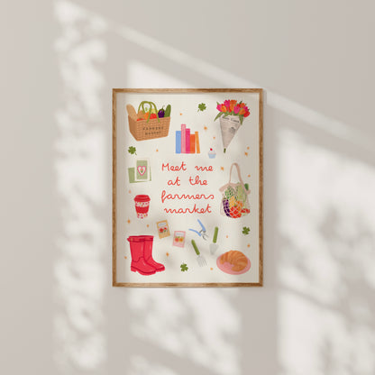 A framed art print featuring a whimsical autumn farmer's market illustration with the text 'Meet me at the farmers market' in red script. The print includes colorful drawings of a basket of vegetables, books, flowers, a tote bag, a red rain boot, gardening tools, and a plate of food, all set against a cream background.