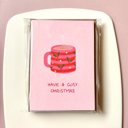 A pink Christmas greeting card in clear cellophane packaging, featuring an illustration of a pink and red striped mug of hot chocolate with holly and stars and the text 'HAVE A COSY CHRISTMAS' on a light pink background.
