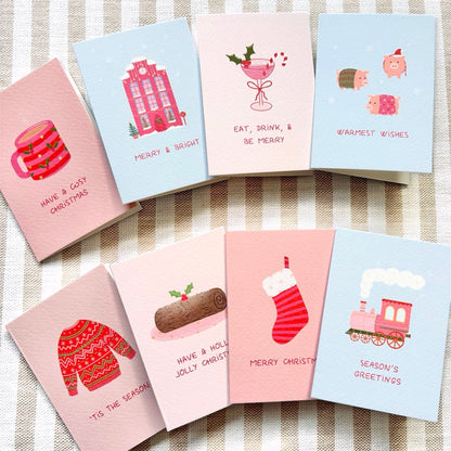 A set of illustrated Christmas greeting cards arranged on a beige-and-white striped fabric background. The cards feature pastel pink and light blue colours with festive whimsical  drawings including pigs in blankets, a snowy pink house, a yule log, and a christmas cocktail, and phrases such as 'Have a cosy Christmas,' 'Merry & Bright,' 'Eat, drink, & be merry,' 'Warmest wishes,' 'Tis the season,' 'Have a holly jolly Christmas,' 'Merry Christmas,' and 'Season's greetings.