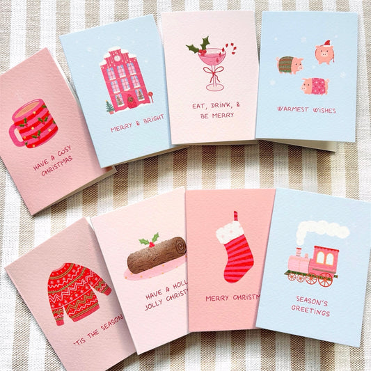 A set of illustrated Christmas greeting cards arranged on a beige-and-white striped fabric background. The cards feature pastel pink and light blue colours with festive whimsical  drawings including pigs in blankets, a snowy pink house, a yule log, and a christmas cocktail, and phrases such as 'Have a cosy Christmas,' 'Merry & Bright,' 'Eat, drink, & be merry,' 'Warmest wishes,' 'Tis the season,' 'Have a holly jolly Christmas,' 'Merry Christmas,' and 'Season's greetings.