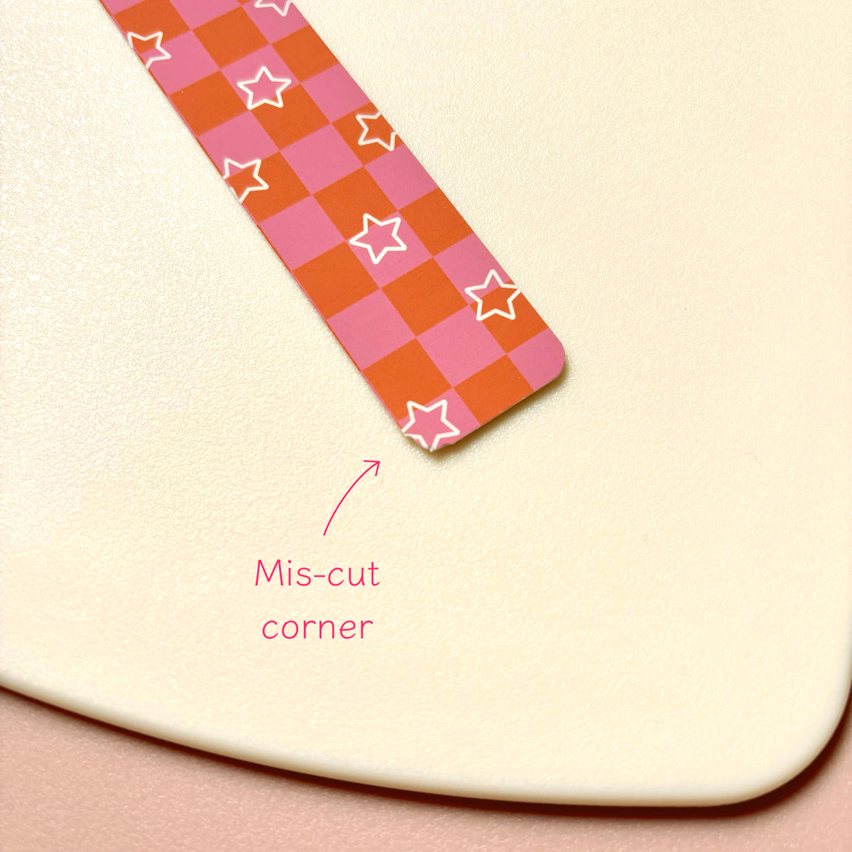 A close-up of a magnetic bookmark with a mis-cut corner, featuring a pink and orange checkerboard patterned strip with white outlined stars. Pink text reads 'Mis-cut corner' with an arrow pointing to the edge.