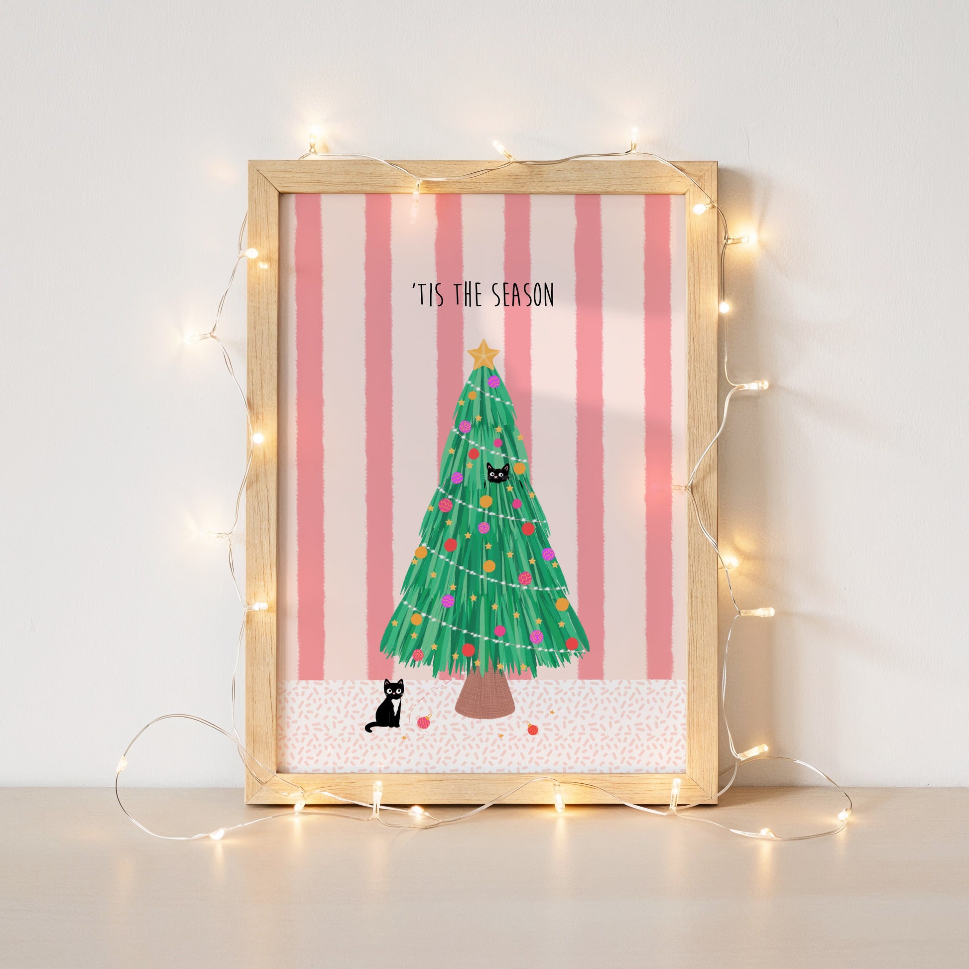 Framed Christmas-themed artwork with a tree and cats on a pink striped background, surrounded by string lights.