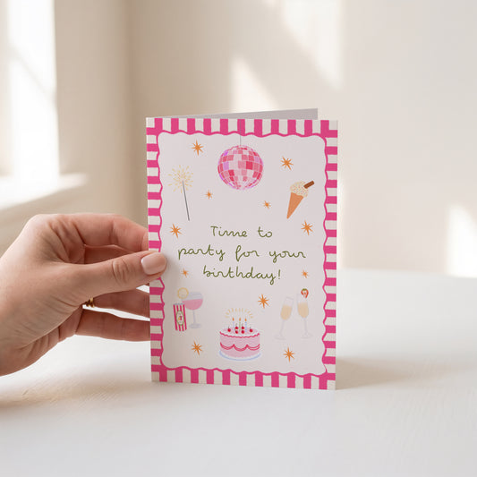 A hand holds a pink and white birthday greeting card with illustrations of a disco ball, cake, champagne, and confetti, against a softly lit indoor background.