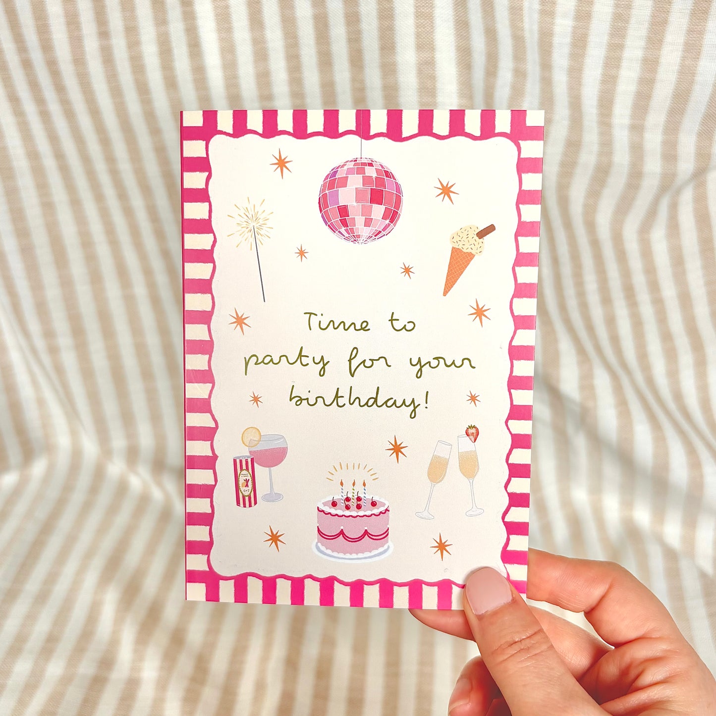 Hand holding a birthday card with pink party-themed illustrations on a striped background