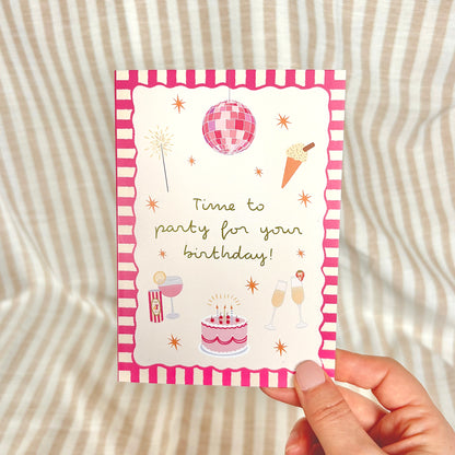 Hand holding a birthday card with pink party-themed illustrations on a striped background