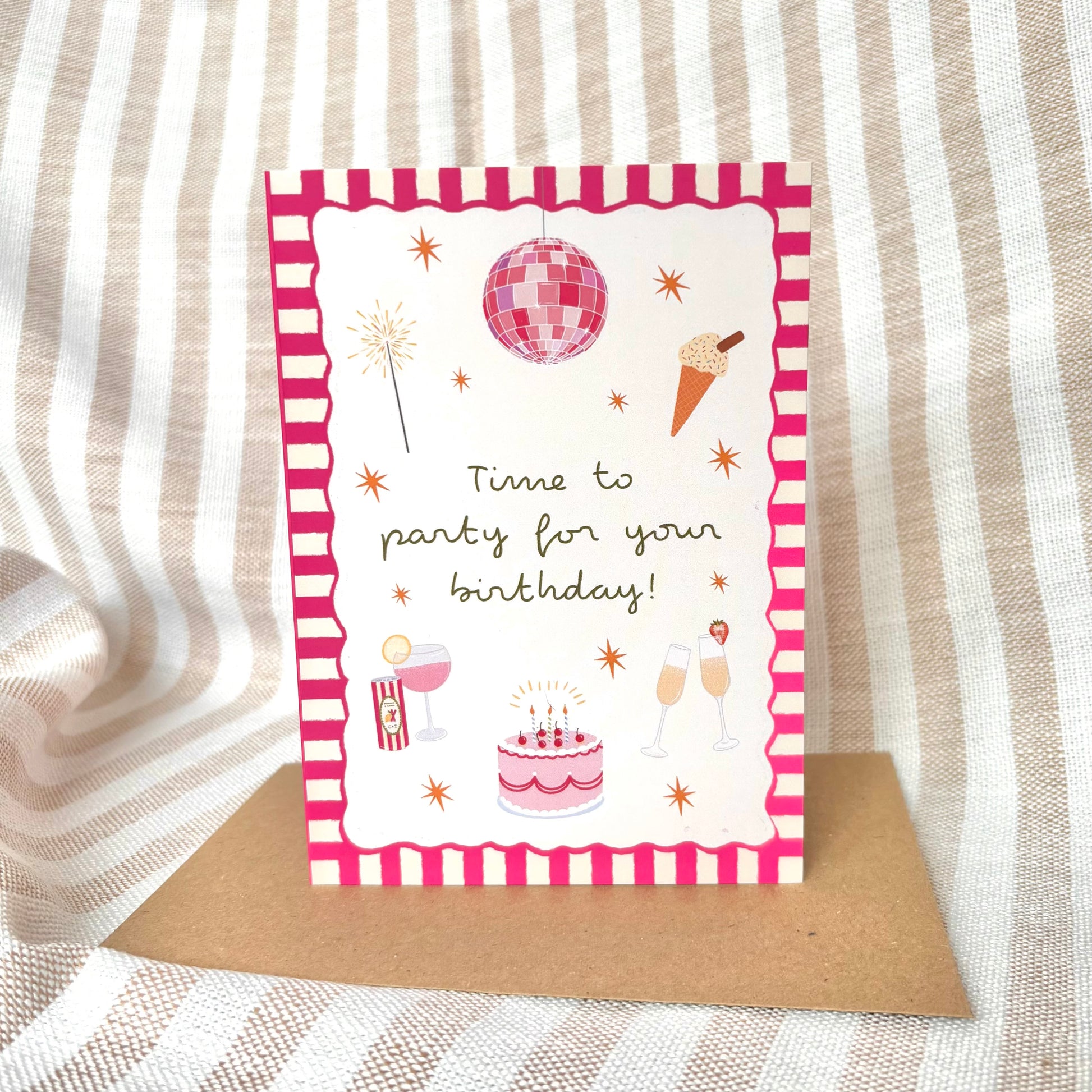 Birthday card with pink party-themed illustrations with a brown kraft envelope on a striped fabric background
