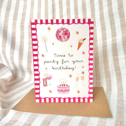 Birthday card with pink party-themed illustrations with a brown kraft envelope on a striped fabric background