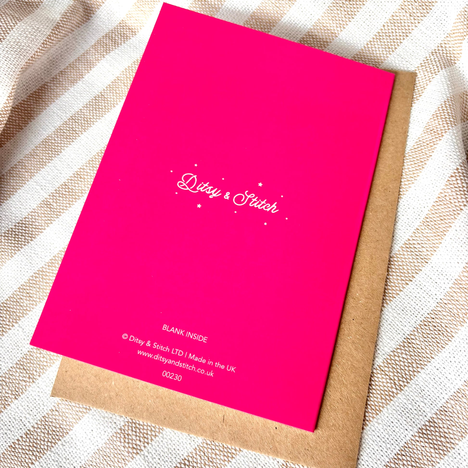 Pink card with 'Ditsy & Stitch' branding on the back, with a brown kraft envelope on a striped fabric background