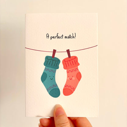 A hand holding a greeting card with a cute illustration of two smiling socks hanging from a clothesline. The card reads 'A perfect match!' in black text.