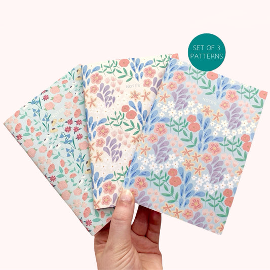 Set of three floral patterned notebooks held by a hand on a white background