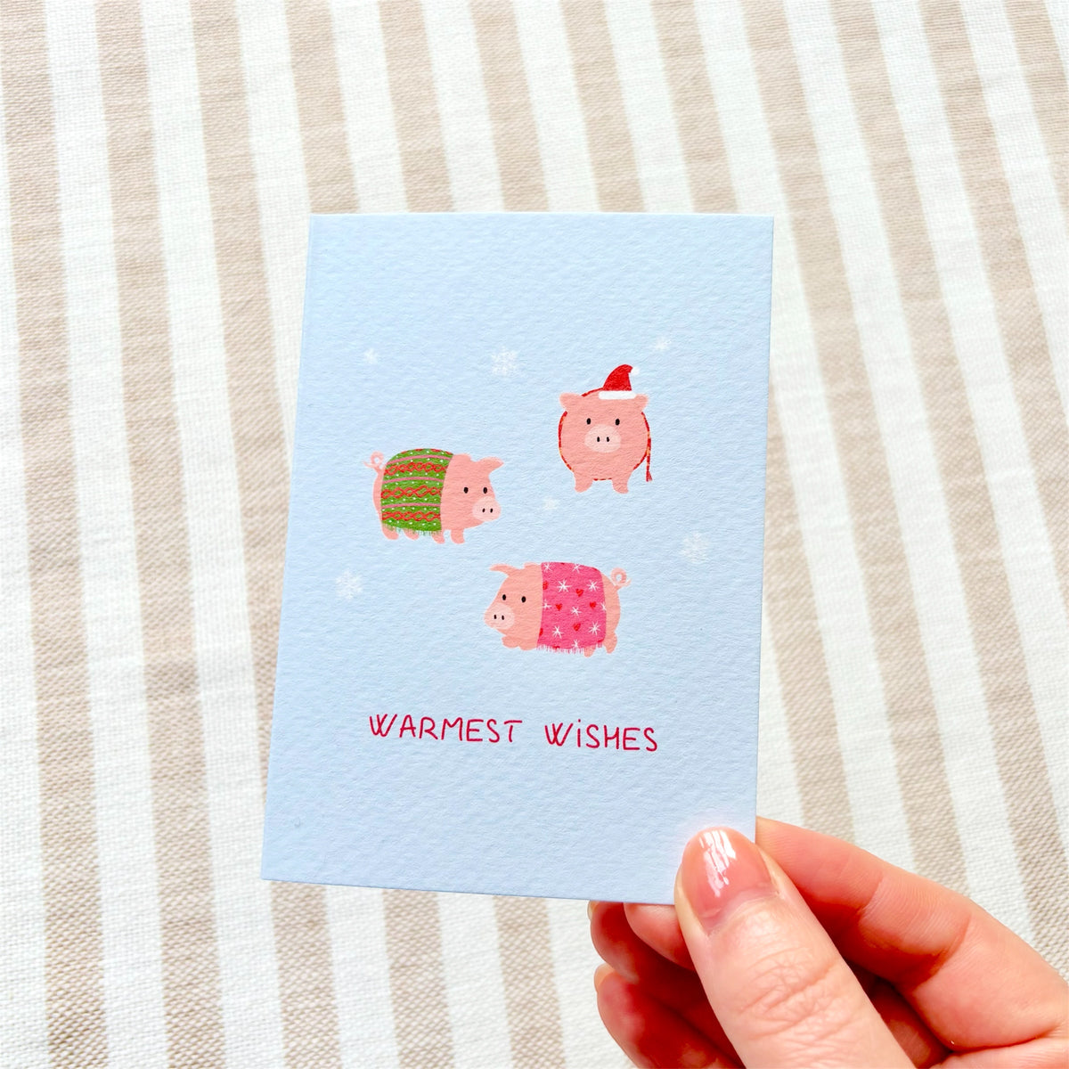 An A7 greeting card with a textured light blue surface, featuring three cute pink pigs wrapped in colourful blankets and wearing hats, with the text 'WARMEST WISHES' in pink at the bottom. The card is photographed at an angle on a soft pastel background.