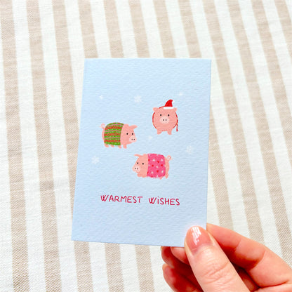 An A7 greeting card with a textured light blue surface, featuring three cute pink pigs wrapped in colourful blankets and wearing hats, with the text 'WARMEST WISHES' in pink at the bottom. The card is photographed at an angle on a soft pastel background.