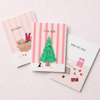 Three illustrated holiday greeting cards arranged on a pale pink background with subtle gold speckles. The center card shows a green Christmas tree with the text "T’IS THE SEASON" and a small black cat beneath it.