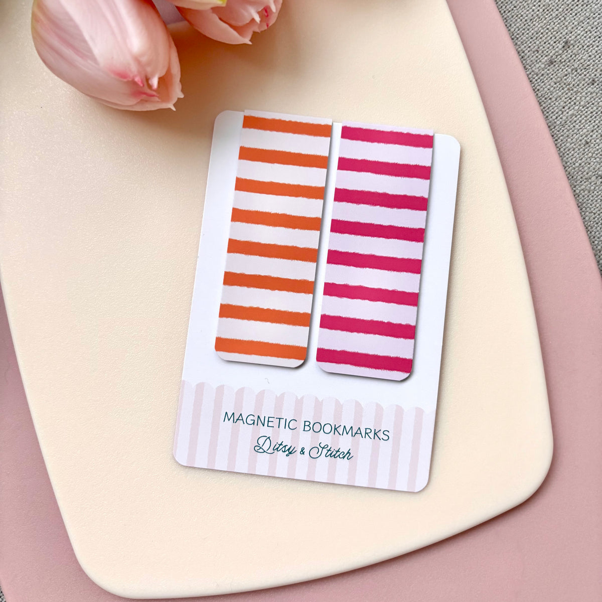 Two striped magnetic bookmarks on a white card labelled 'MAGNETIC BOOKMARKS Ditsy & Stitch', placed on a beige background with pink tulips in the corner.