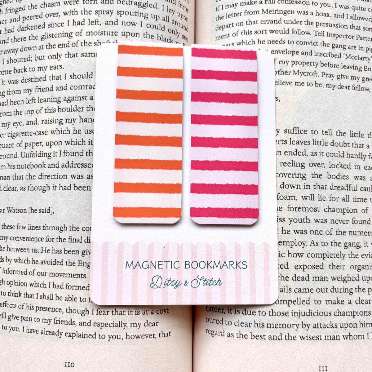 Two rectangular magnetic bookmarks with rounded corners in warm orange and pink tones, displayed on an open book page with printed text.