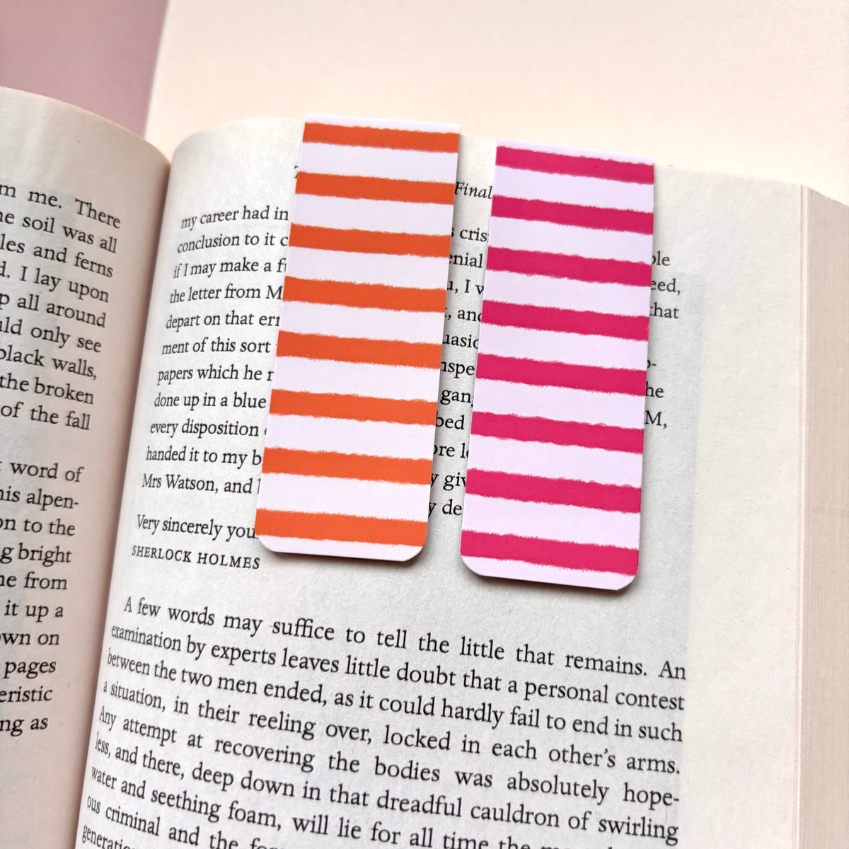 Two striped paper bookmarks with orange and white stripes on the left and pink and white stripes on the right, placed on open book pages.