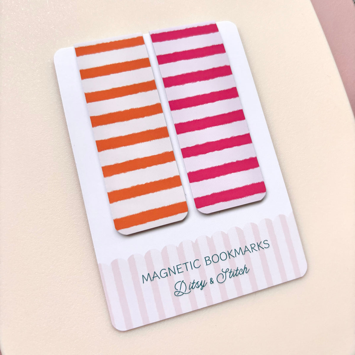 Two striped magnetic bookmarks on a white display card. One bookmark has orange and white stripes, and the other has pink and white stripes.