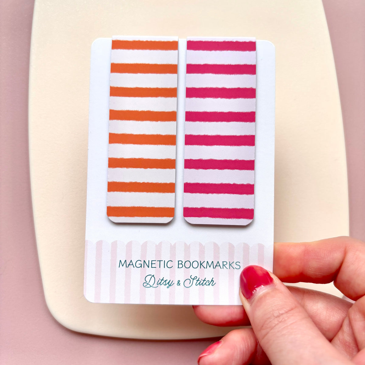 A hand with red nail polish holds a white card displaying two striped magnetic bookmarks, one with orange and white stripes and the other with pink and white stripes. The card reads 'MAGNETIC BOOKMARKs Ditsy & Stitch'.