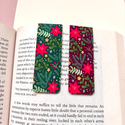 Two rectangular bookmarks with a festive Christmas botanical pattern, resting on open book pages. The left bookmark has a dark green background and the right has a dark purple background, both are patterned with red poinsettias, green leaves and stars.