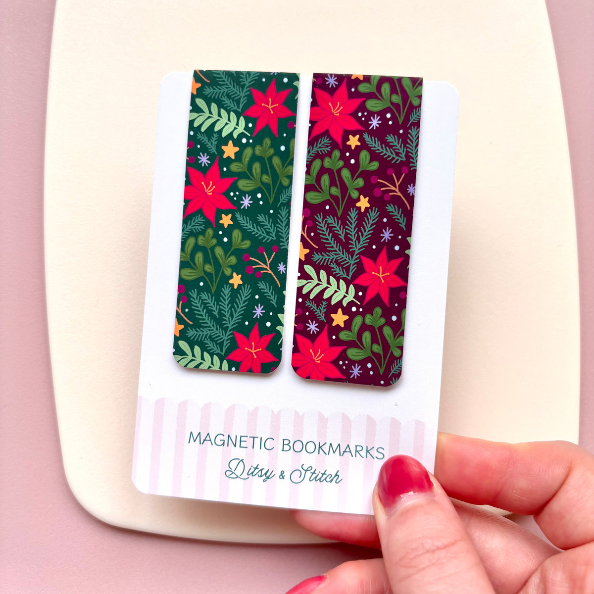 A hand with red nail polish holds a white card displaying two rectangular magnetic bookmarks with a festive Christmas pattern. The bookmarks feature red poinsettias, green pine branches, and small yellow stars on dark green and purple backgrounds.
