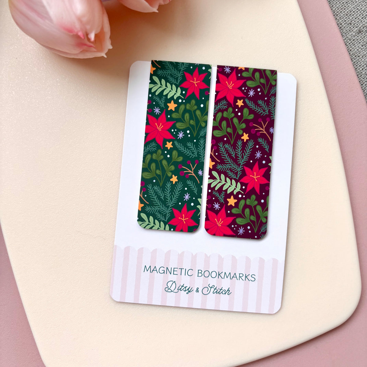 Two rectangular magnetic bookmarks with festive Christmas patterns, displayed on a white card labelled 'MAGNETIC BOOKMARKS' by 'Ditsy & Stitch'. The bookmarks feature holiday motifs including poinsettias, pinecones, and stars in green, red, and dark purple tones.
