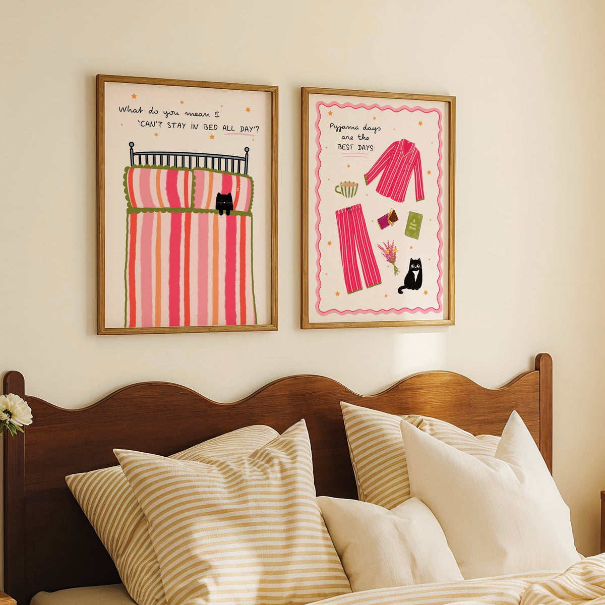 Two framed art prints hanging on a cream wall above a wooden bed with striped pillows. The left print shows a striped bed with the text 'What do you mean I CAN'T STAY IN BED ALL DAY?'’. The right shows illustrated pyjamas, book, hot chocolate and  black cat with the text ‘Pyjama days are the BEST DAYS’.