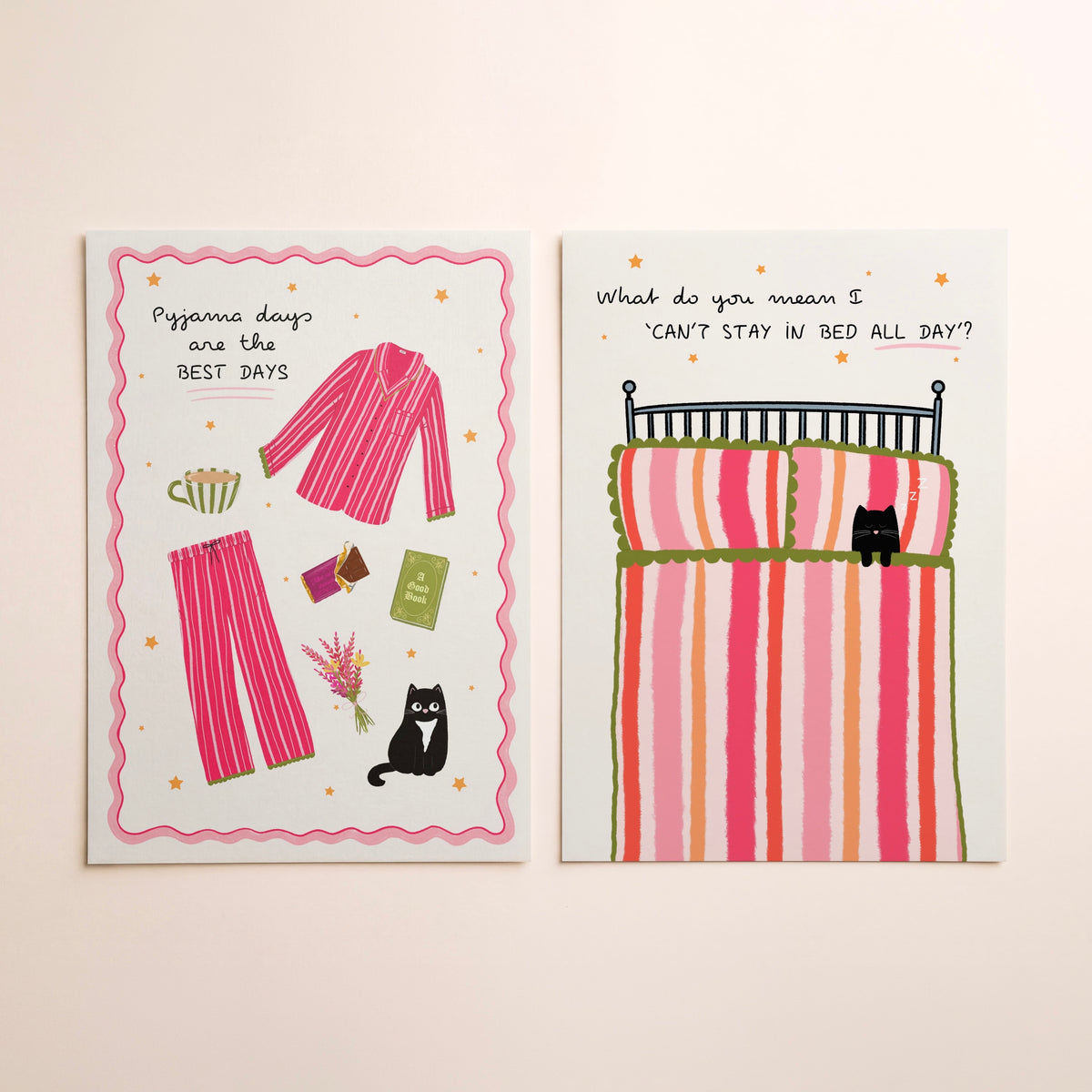 Two art prints with whimsical illustrations lying side-by-side on a warm beige background. The left print shows pink striped pyjamas and a black cat with the text 'Pyjama days are the BEST DAYS'. The right shows a bed with a striped duvet and a cat, with text saying ‘What do you mean I can’t stay in bed all day?’.