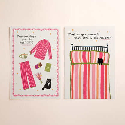 Two art prints with whimsical illustrations lying side-by-side on a warm beige background. The left print shows pink striped pyjamas and a black cat with the text 'Pyjama days are the BEST DAYS'. The right shows a bed with a striped duvet and a cat, with text saying ‘What do you mean I can’t stay in bed all day?’.