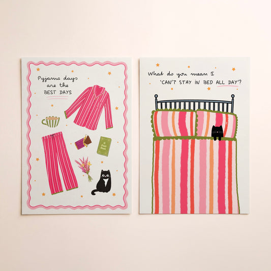 Two art prints with whimsical illustrations lying side-by-side on a warm beige background. The left print shows pink striped pyjamas and a black cat with the text 'Pyjama days are the BEST DAYS'. The right shows a bed with a striped duvet and a cat, with text saying ‘What do you mean I can’t stay in bed all day?’.