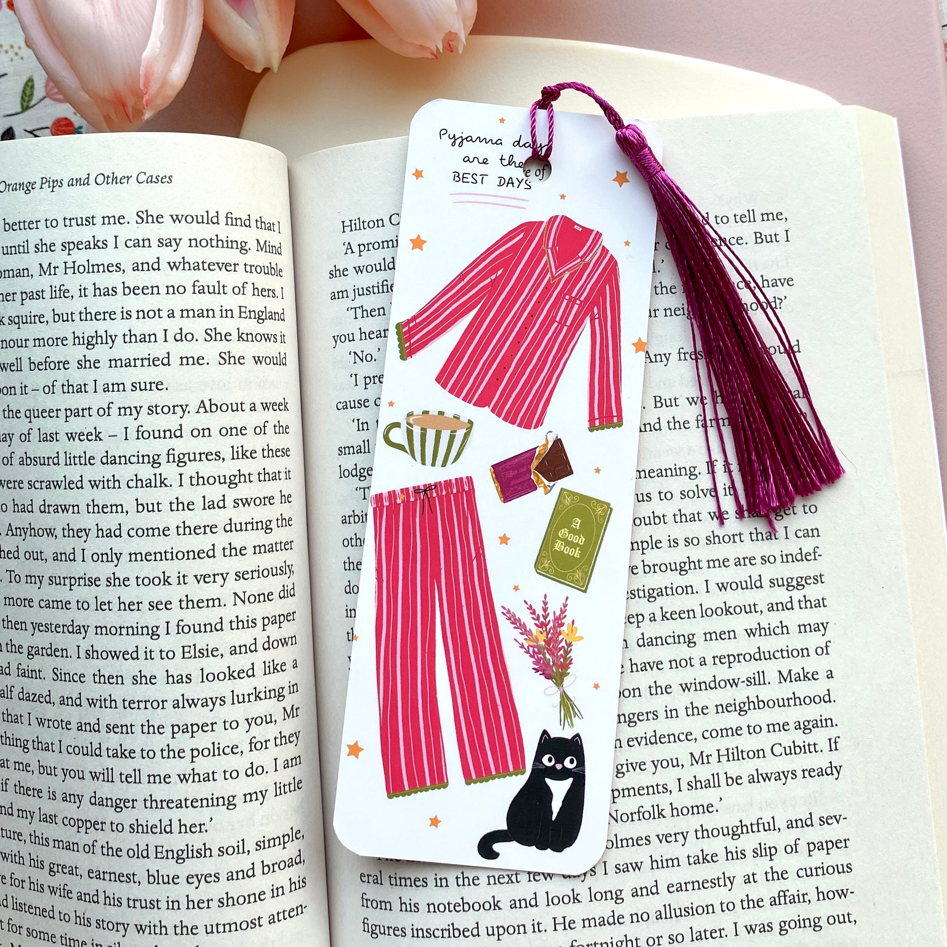 Bookmark with illustrations of dark pink striped pyjamas, black cat, and other items with a purple tassel on an open book.