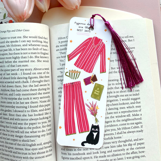 Bookmark with illustrations of dark pink striped pyjamas, black cat, and other items with a purple tassel on an open book.