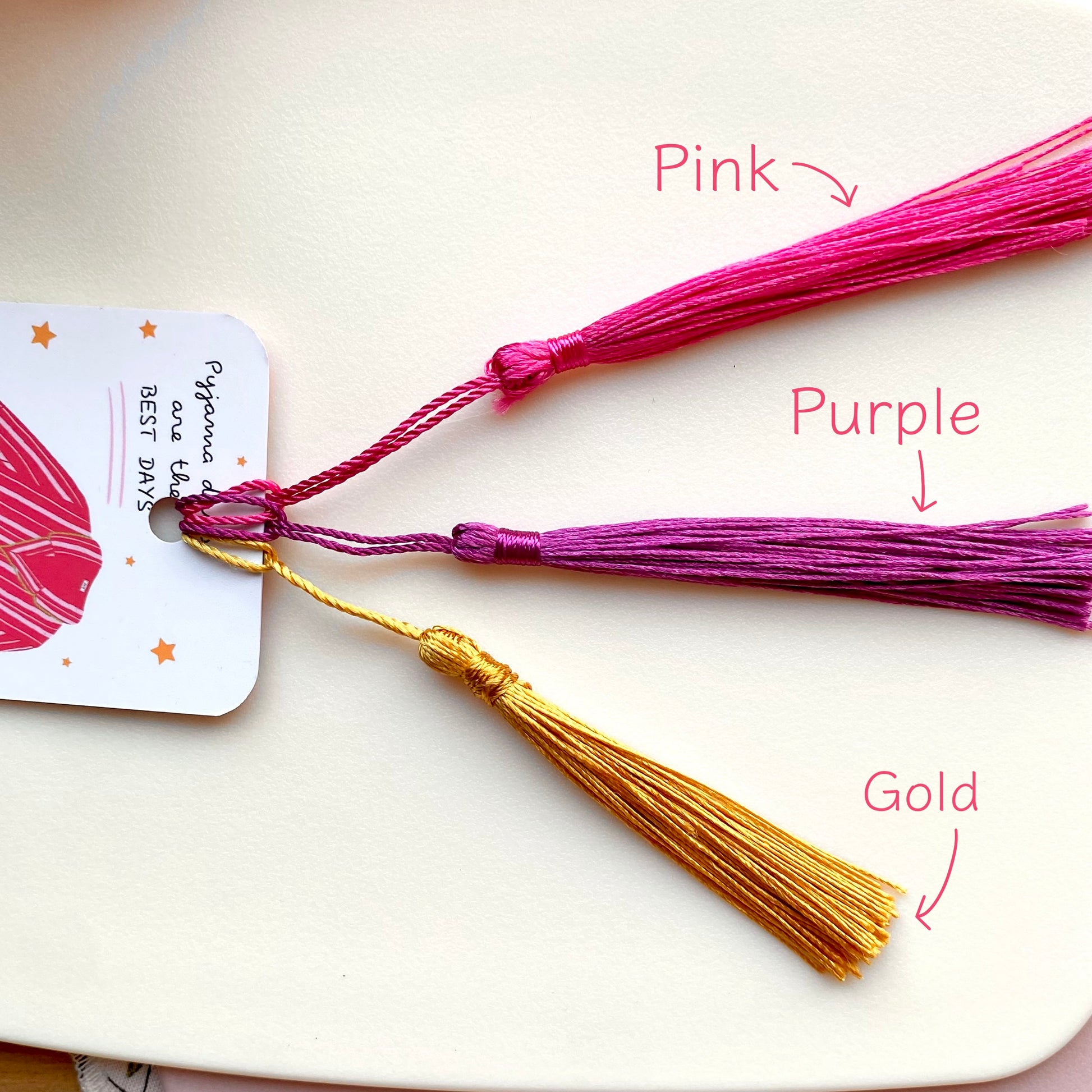 Three tassels in pink, purple, and gold on a white surface with labels indicating their colors.