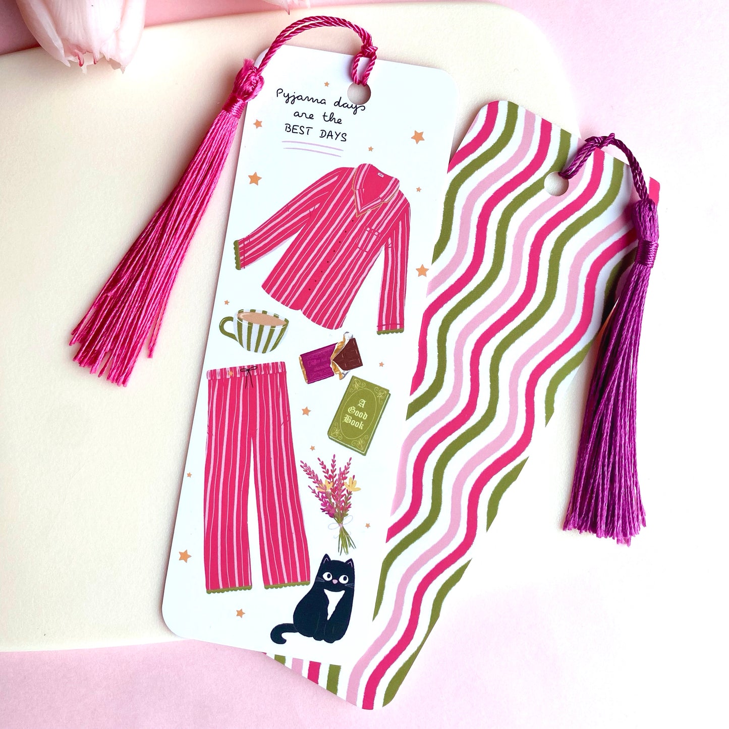 Bookmark with pink tassels featuring illustrations of pyjamas, a cat, a book, chocolate, and scattered stars, with pink and green wavy stripes on the reverse side, on a pink background.