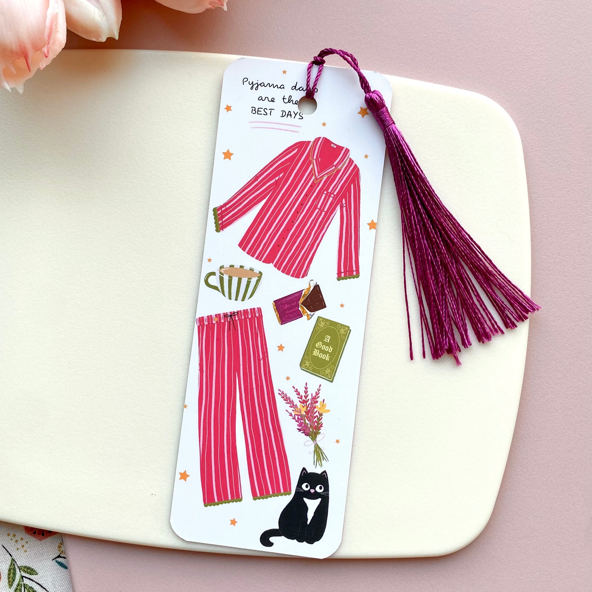 Bookmark with pink striped pyjama design with a cat, green book, green striped mug and stars, with tassel on a white surface