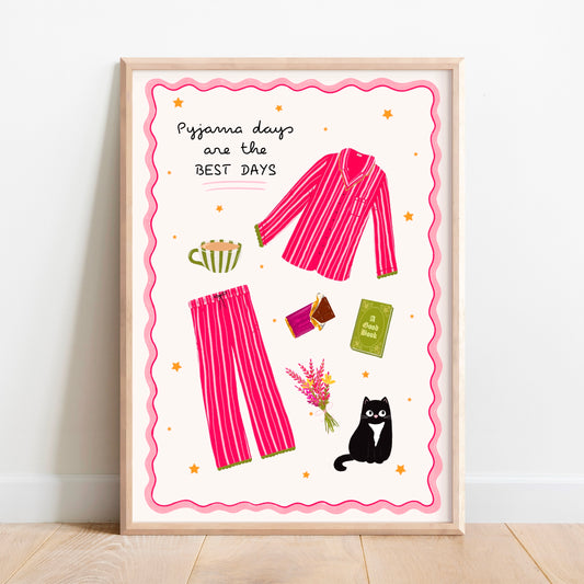 A framed art print leaning against a white wall on a light wood floor. The print features a whimsical illustration of pink striped pajamas, a mug, a book, flowers, and a black cat with the text 'Pyjama days are the BEST DAYS' on a cream background with a pink wavy border.
