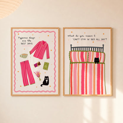 Two framed art prints with whimsical illustrations hang side-by-side on a warm beige wall. The left print shows pink striped pyjamas and a black cat with the text 'Pyjama days are the BEST DAYS'. The right shows a bed with a striped duvet and a cat, with text saying ‘What do you mean I can’t stay in bed all day?’.