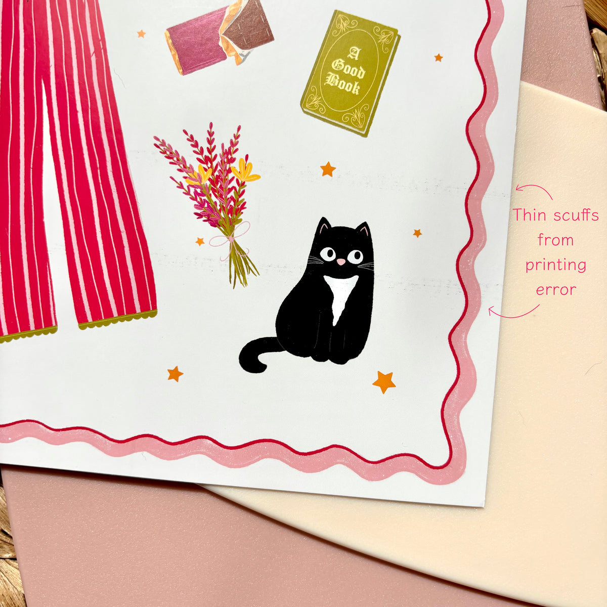 A close-up of a printed art print showing a whimsical illustration: a black cat with a white chest, pink curtains, flowers, stars, and a small book. A pink handwritten note reads 'Thin scuffs from printing error.