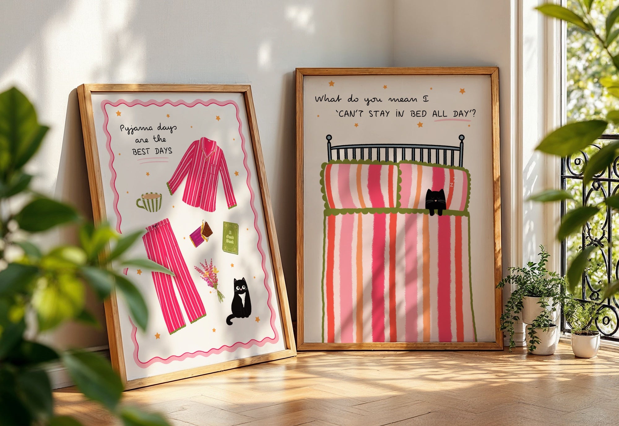 Two framed pyjama day-themed illustrations with pink beds and black cats, placed on a wooden floor and surrounded by houseplants.