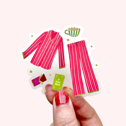 Hand holding a transparent vinyl sticker with pink pyjamas and green and purple cosy day accessories on a white background