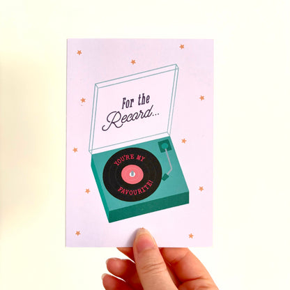 A hand holding a greeting card with a retro record player illustration. The card has a lilac background with small gold stars and the text 'For the Record… you’re my favourite’.