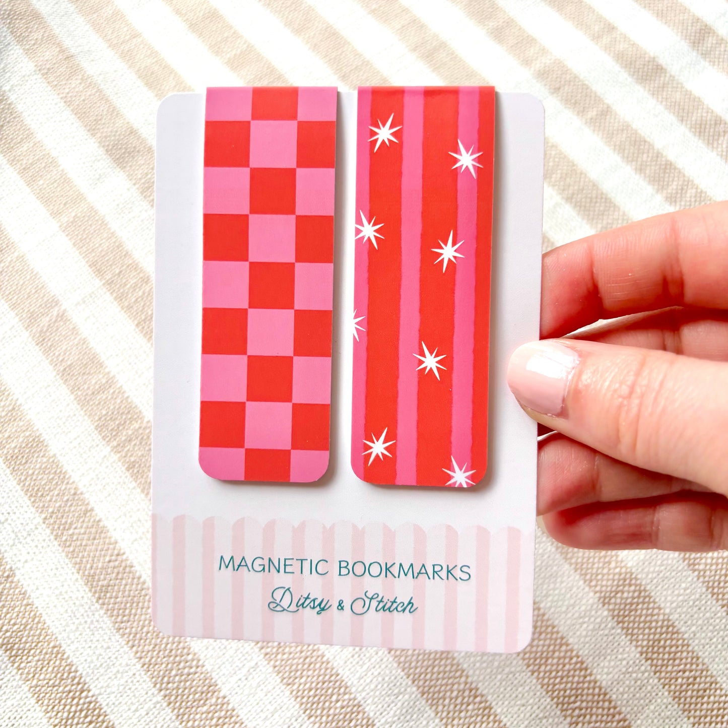 Two pink and red patterned magnetic bookmarks on a card held by a hand against a striped background