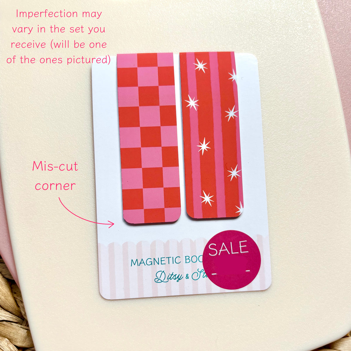 A close-up of a magnetic bookmark set with two decorative patterned bookmarks: one red and pink checkerboard design and one pink and red vertical stripe design with white starbursts. Pink text on the card notes that imperfections may vary in the set and points out a mis-cut corner, with a SALE sticker overlay.