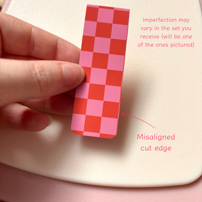 A hand holds a magnetic bookmark with a pink and red checkerboard pattern against a light cream background, with pink text noting that imperfections may vary and pointing out a misaligned cut edge.